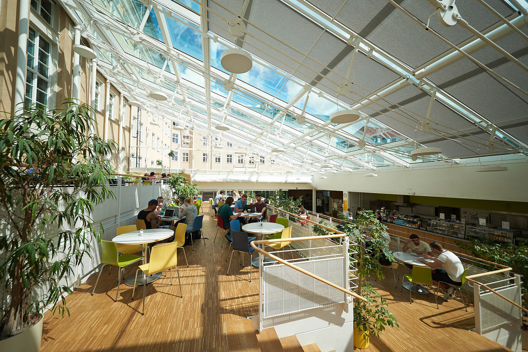 HTWK Leipzig Mensa HTWK Leipzig Mensa Innenansicht. Auf einem holzfarbenen Boden stehen Tische und Stühle an denen Menschen essen. Die Decke ist schrägt geneigt mit großen Fenstern durch die viel Licht einfällt. In mehreren Blumentöpfen stehen Pflanzen.