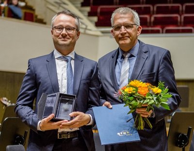 Karsten Petrusch überreicht Dr.-Ing. Andreas Reinhold den Dissertationspreis 2018 der Stiftung HTWK. Übergabe des Dissertationspreises 2019. Zwei Menschen in Anzug halten die Urkunde, den Preis und einen Blumenstrauß in den Händen.