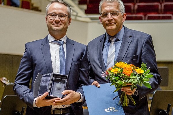 Übergabe des Dissertationspreises 2019. Zwei Menschen in Anzug halten die Urkunde, den Preis und einen Blumenstrauß in den Händen.