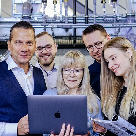 Sechs Personen stehen dicht zusammen, blicken auf einen geöffneten Laptop und ein Informationsblatt, im Hintergrund sichtbare Rohrleitungen und Technik.