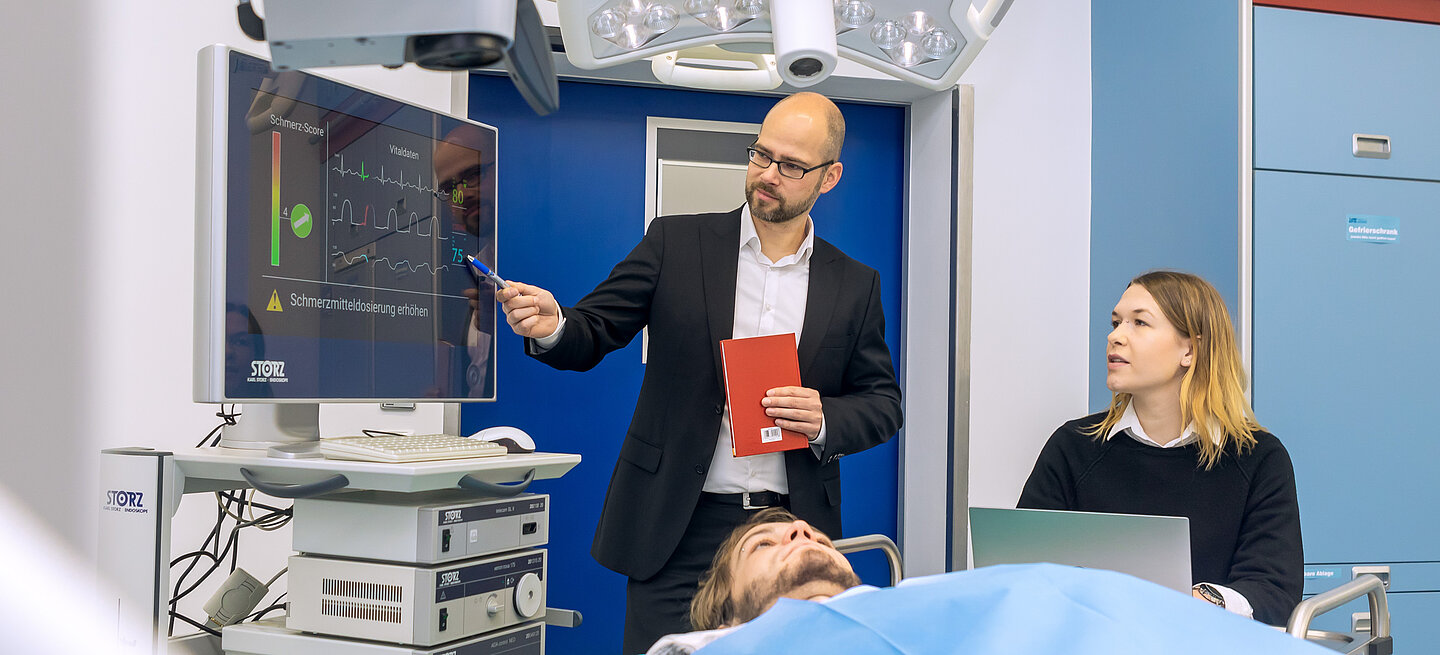 Ein Mann erklärt Biosignale auf einem Monitor, während eine Frau daneben sitzt und eine Person im Bett liegt