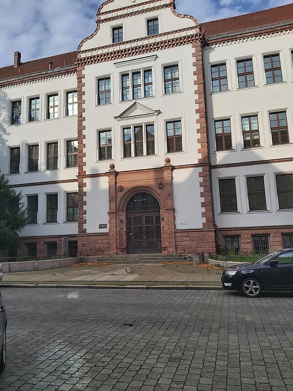 Das Bild zeigt die Fassade eines historischen Gebäudes, das mit seinem prägnanten Eingangsbereich und seiner charakteristischen Architektur ins Auge fällt. Die Fassade ist in weißem Putz gehalten, während die Fensterrahmen und Ecken durch rotbraunen Stein hervorgehoben werden. Der Haupteingang ist kunstvoll gestaltet, mit einem großen, verzierten Rundbogen aus roten Steinen und dekorativen Elementen. Darüber hinaus sorgen symmetrisch angeordnete Fenster und der zentrale Giebel dafür, dass das Gebäude klassisch und zugleich beeindruckend wirkt.