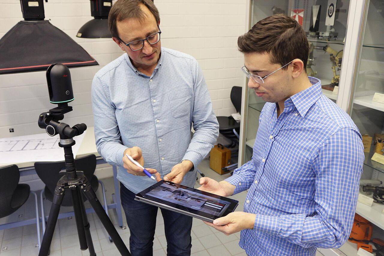Holger Evers und Christian Musella stehen in einem Labor und schauen gemeinsam auf ein Tablet. Links im Bild steht auf einem Stativ ein Laserscanner im Raum. Mit dem Hightech-Gerät arbeitet Musella im Rahmen seiner Doktorarbeit.
