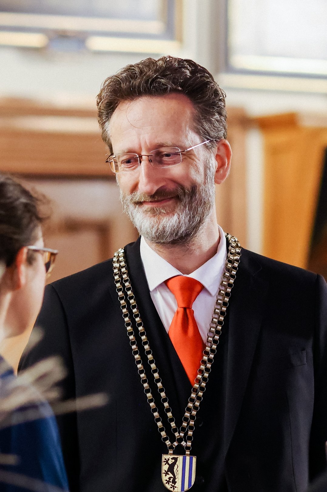 Mann in schwarzem Anzug mit orangefarbener Krawatte und einer großen Kette mit Wappen um den Hals steht in einem Raum und spricht mit einer Frau mit Brille und dunklen Haaren