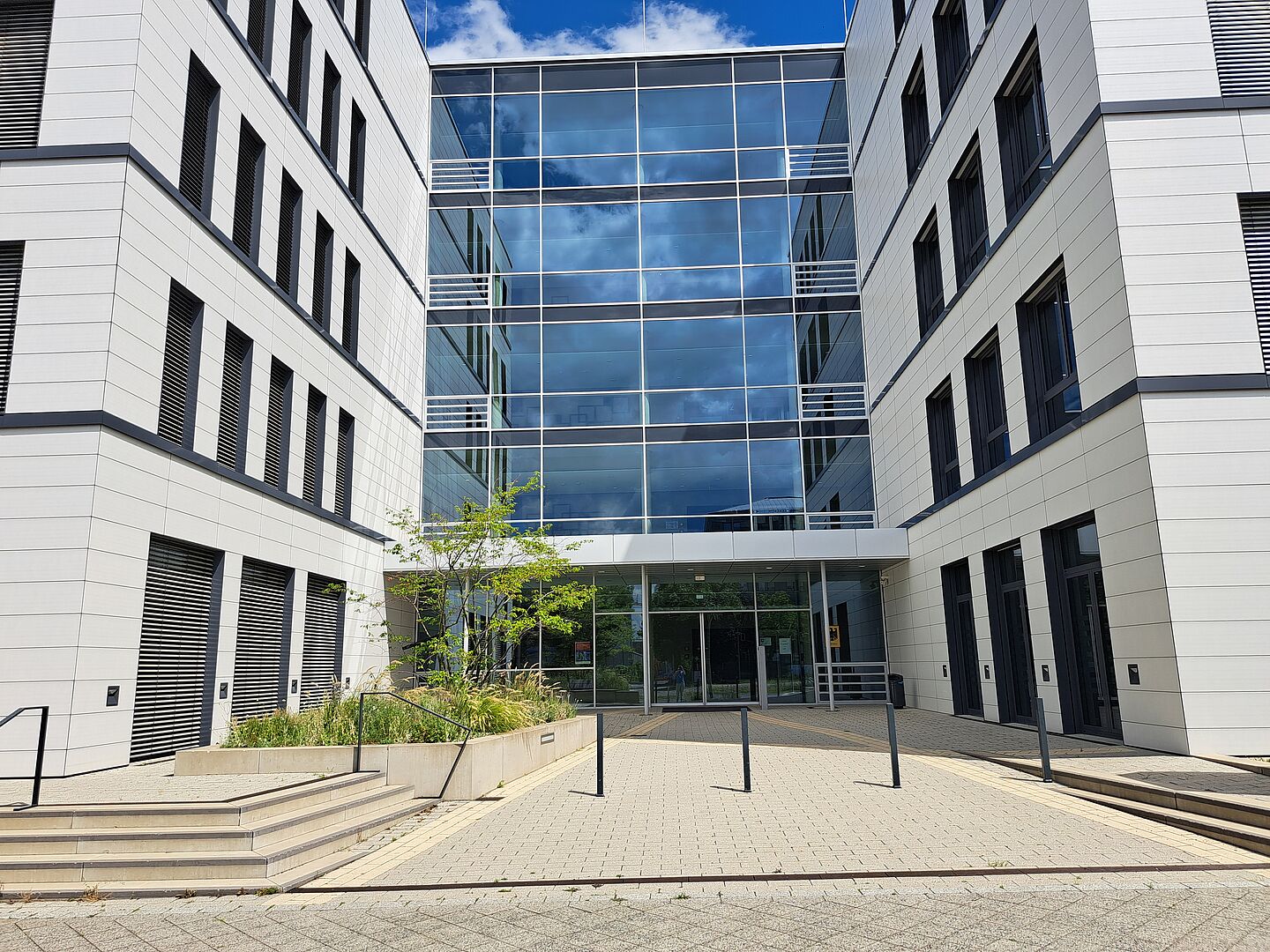 Das Bild zeigt das Bundesamt für Naturschutz, ein modernes Bürogebäude mit einer Kombination aus glasigen und weißen Fassadenelementen. Die Architektur ist symmetrisch gestaltet, mit einem zentralen Glasportal, das als Haupteingang dient. Dieser Bereich ist durch eine transparente Überdachung geschützt. Zu beiden Seiten des Glaszentrums erstrecken sich weiße Gebäudeflügel mit vertikal ausgerichteten, schwarzen Fensterläden. Vor dem Gebäude führt ein gepflasterter Weg zum Eingang, flankiert von bepflanzten Bereichen und jungen Bäumen.