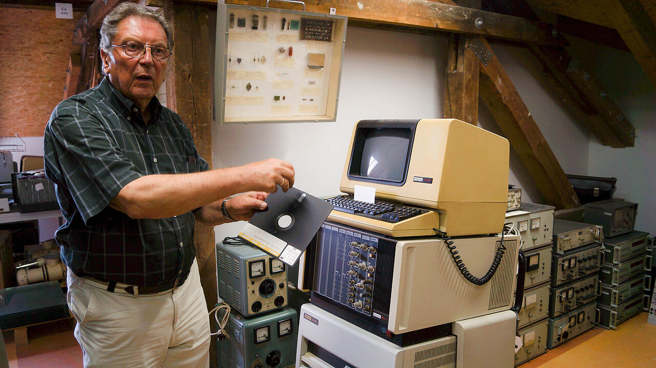 Ein Mann steht vor einem sehr alten Computer und hält eine Diskette in der Hand.