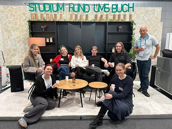 Studierende und Mitarbeiter der HTWK sitzen und posieren freudig auf der Bühne des Messestandes. 