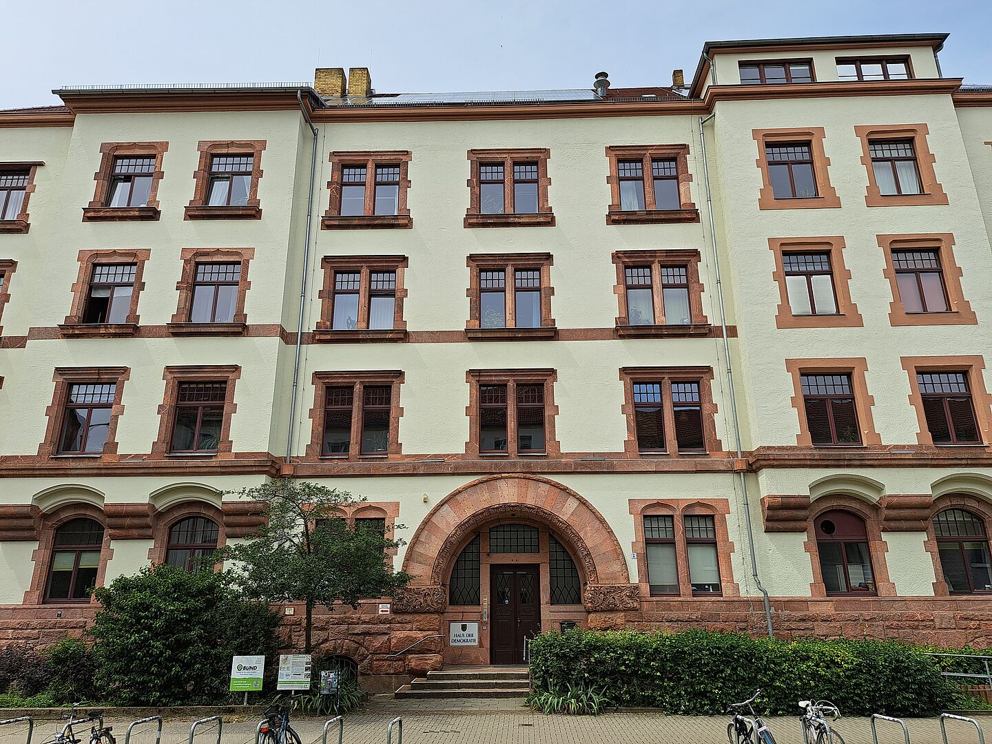  Das Bild zeigt das Haus der Demokratie. Es handelt sich um ein mehrstöckiges Gebäude mit einer cremefarbenen Fassade und rotbraunen Fensterrahmen sowie Verzierungen um die Fenster. Der Haupteingang ist von einem großen, rundbogigen Portal umgeben. Vor dem Gebäude befinden sich Fahrradständer und einige Büsche.