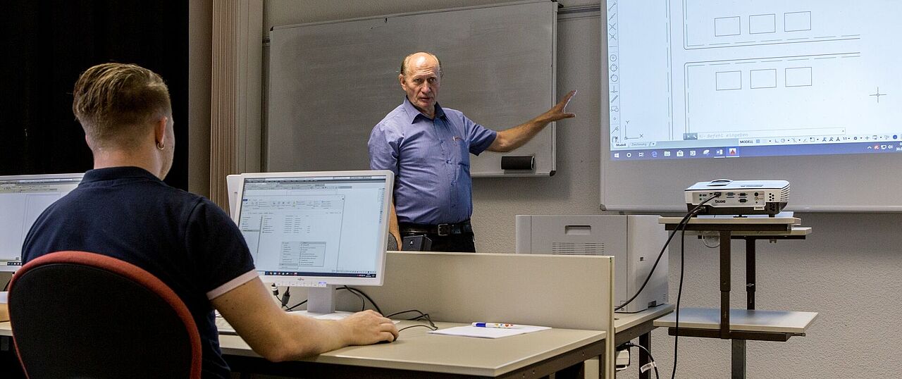 Ein Dozent steht in einem Seminarraum vorn und zeigt auf eine Präsentation. Ein Lehrgangsteilnehmer sitzt mit dem Rücken zur Kamera vor einem Computer.