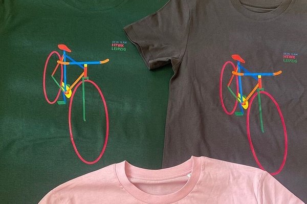  Drei T-Shirts in Pink, Grau und Grün mit identischem buntem Fahrradaufdruck liegen nebeneinander auf einer grünen Fläche.
