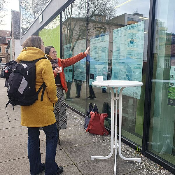 Frau erklärkt anderer Frau Poster, das hinter einer Scheibe hängt.