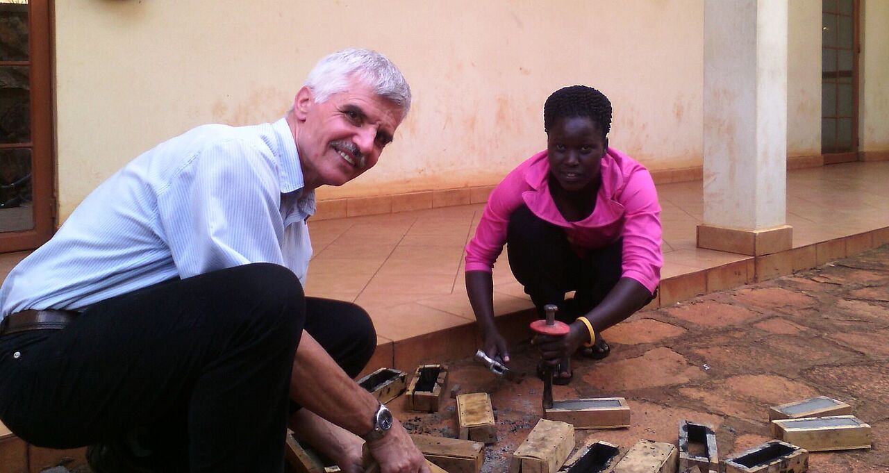 In einem Hof bearbeiten Prof. Klaus Gaber und eine dunkelhäutige Studentin am Boden kniend Werkstücke. 