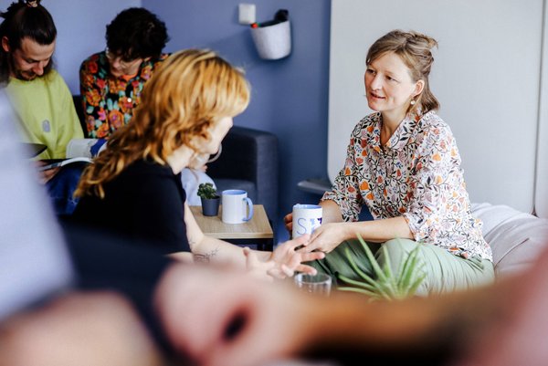 Mehrere Personen sitzen in einem Besprechungsrauum. Zwei Frauen im Vordergrund unterhalten sich, im Hintergrund lesen zwei Personen ein Buch.
