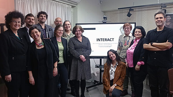 Gruppenbild INTERACT Das Foto zeigt 12 Frauen und Männer stehend vor einem Bildschirm mit der Aufschrift INTERACT