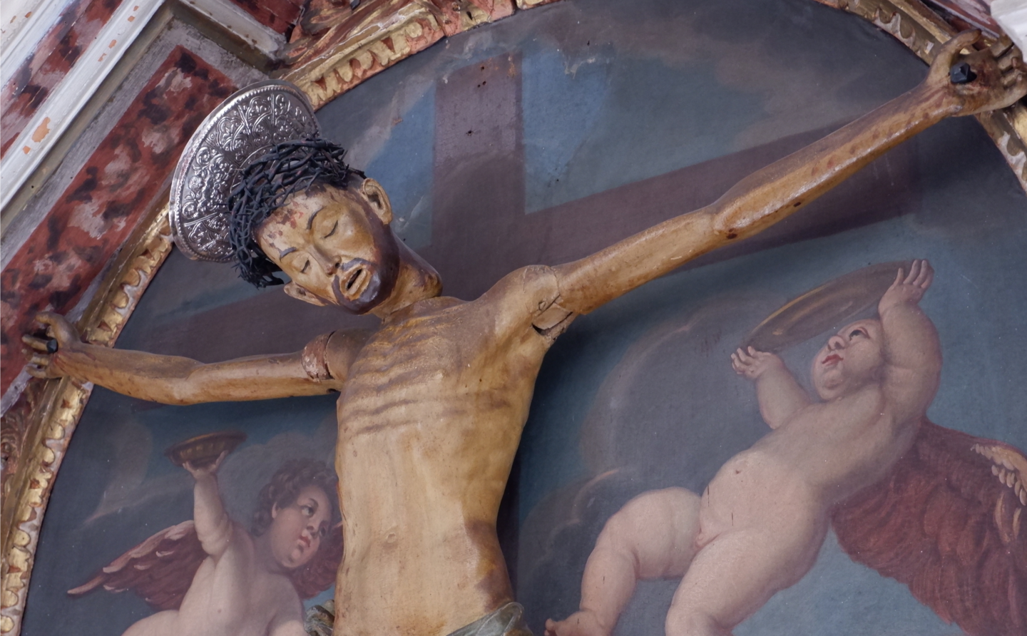 Holzfigur des gekreuzigten Christus mit beweglichen Armen und Kopf, vor einem Gemälde mit zwei Engeln und einem Kreuz