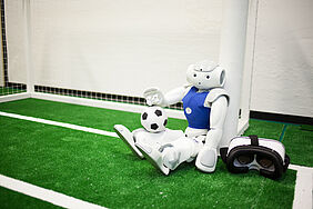 Nao-Roboter „Gargamel“ sitzt an einem Tor mit Fußball und VR-Brille daneben.