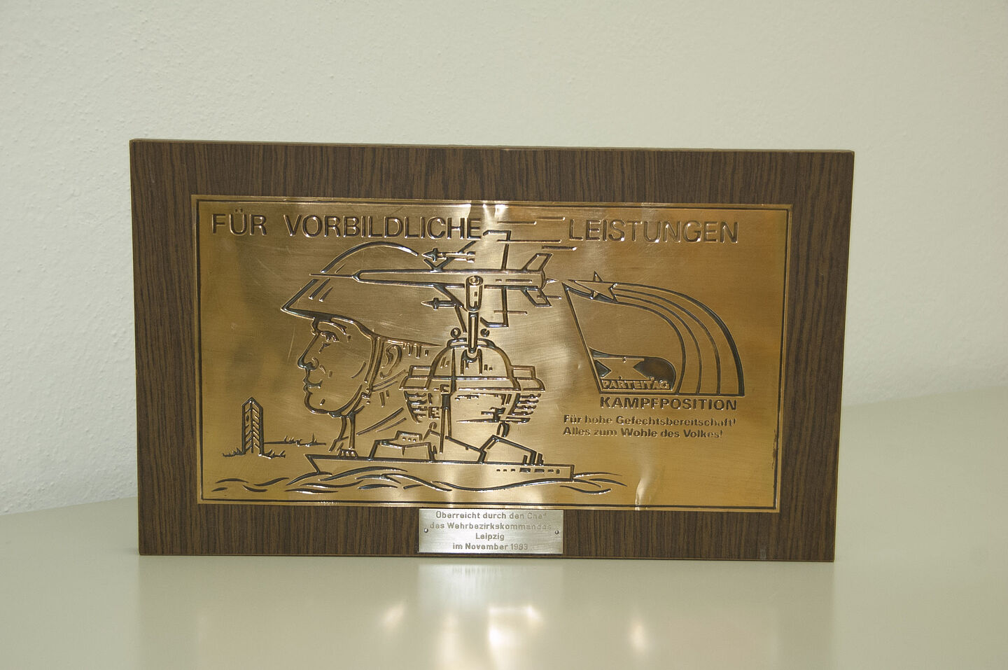 Ehrentafel aus dem HTWK-Archiv (Foto: HTWK Leipzig) Zu sehen ist eine Ehrentafel aus Messing in einem braunen Holzrahmen. Auf der Tafel steht eingefräst: "Für vorbildliche Leistungen. Für hohe Gefechtsbereitschaft. Alles zum Wohle des Volkes." Die Tafel ist 1983 durch das Wehrbezirkskommando Leipzig überreicht worden.