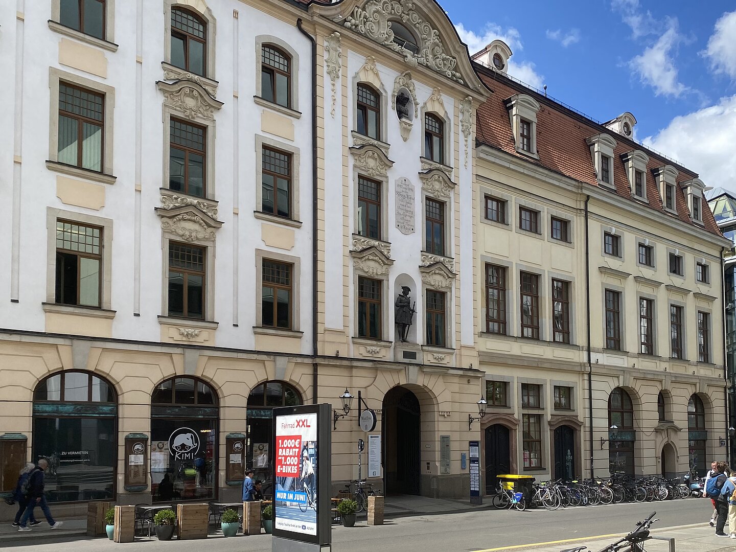 UBL Musik Das Bild zeigt eine Straßenszene mit mehreren historischen Gebäuden, die reich an architektonischen Verzierungen sind. Die Fassaden sind eine Mischung aus Weiß und Beige mit großen Fenstern und dekorativen Stuckarbeiten. Ein Gebäude hat eine Statue in einer Nische über dem Eingang. Entlang des Bürgersteigs sind Fahrräder geparkt, und es sind einige Menschen zu Fuß unterwegs. Im Vordergrund befindet sich ein digitales Werbeschild.