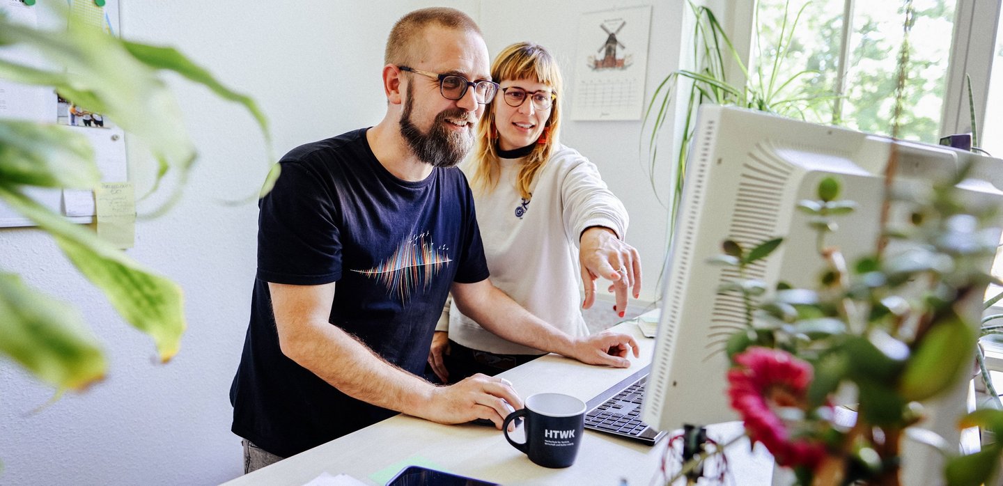 Ein Mann und eine Frau stehen an einem Schreibtisch und schauen auf einen Computerbildschirm. Die Frau zeigt auf den Bildschirm, beide wirken konzentriert. Im Vordergrund sind Pflanzen und Büromaterialien zu sehen.
