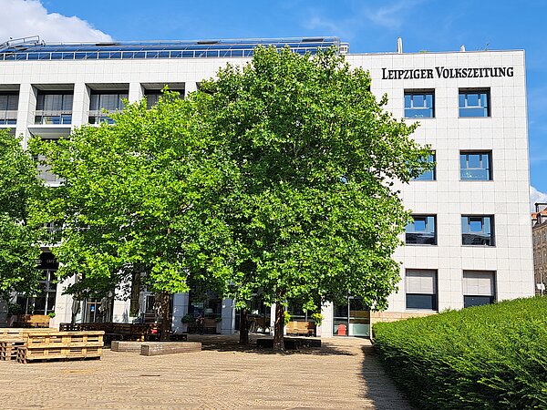 Ein weißes Gebäude mit Solaranlage auf dem Dach. Oben an dem Gebäude steht in Großbuchstaben „Leipziger Volkszeitung“. Vor dem Gebäude sind einige Bänke und grüne Bäume.