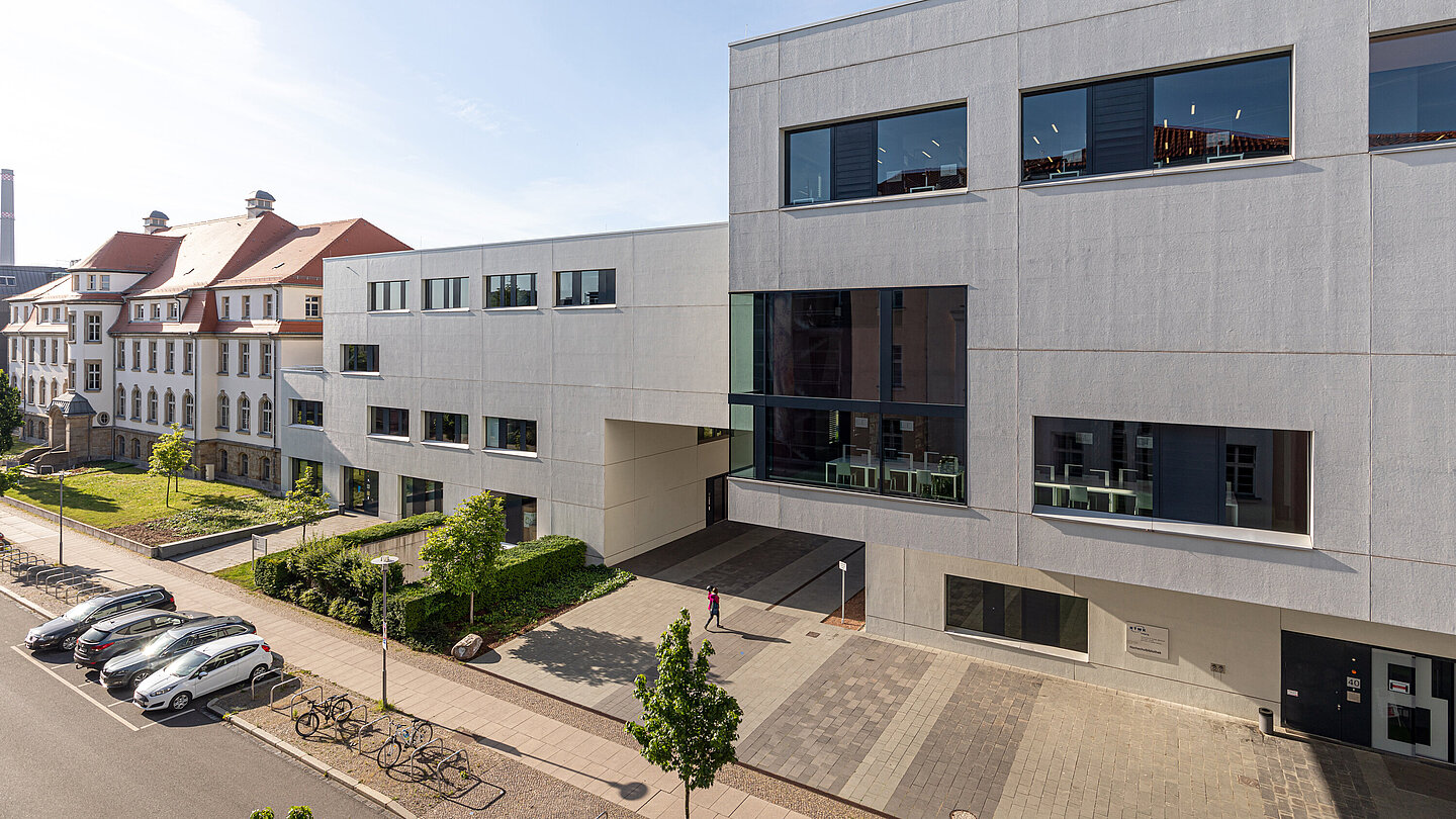 Im Vordergrund zwei weiße, moderne kubische Gebäude an einer Straße. Im Hintergrund ein älteres Hochschulgebäude.