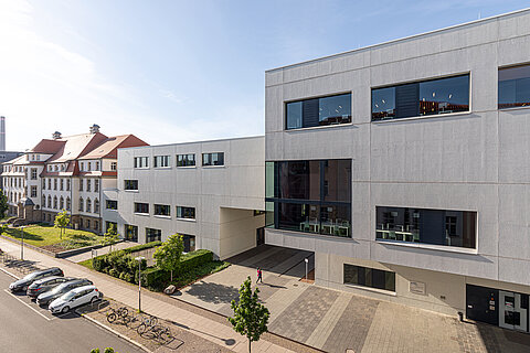 Hochschulbibliothek und Medienzentrum Im Vordergrund zwei weiße, moderne kubische Gebäude an einer Straße. Im Hintergrund ein älteres Hochschulgebäude.