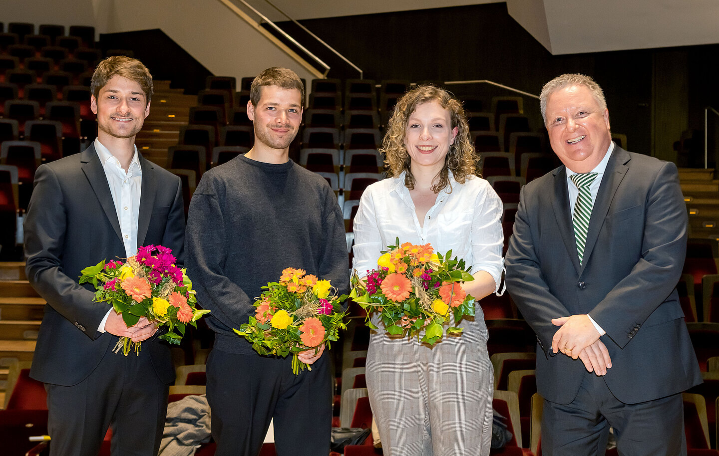 Zwei Männer und eine Frau je mit Blumenstrauß in der Hand sowie ein weiterer Mann stehen auf der Bühne im großen Saal des Gewandhauses