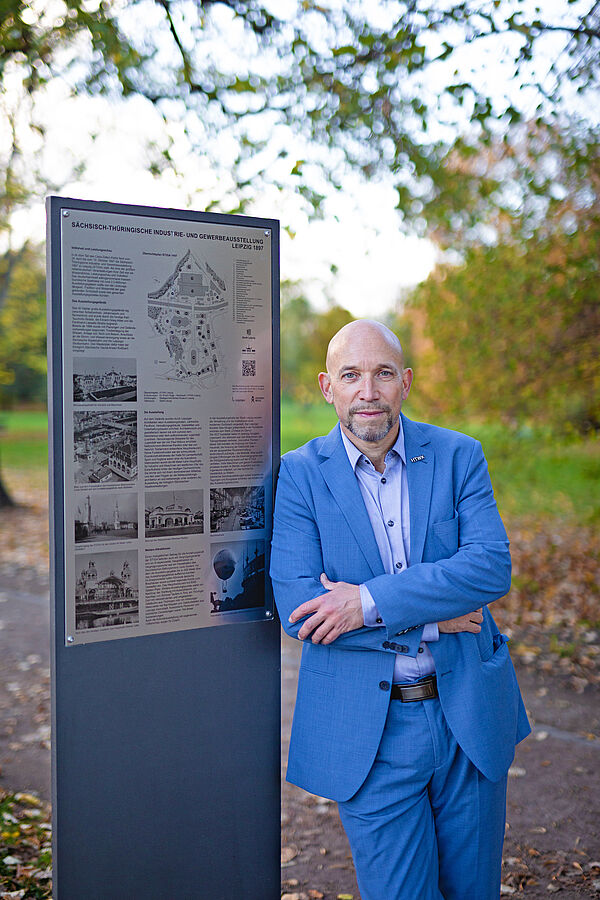 Dr. Ruge steht im blauen Anzug neben einer Stele im Clara-Zetkin-Park