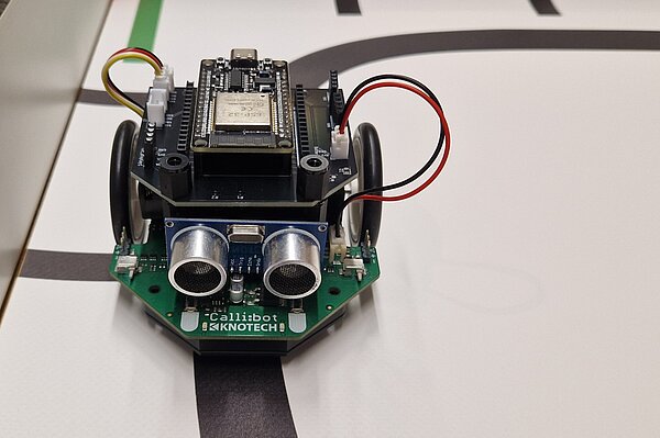 CalliBot 2 mit ESP32 ähnliche einem Miniaturauto auf einem Mikrocontroller mit Kameraaugen und Kabeln, fährt auf einer schwarzen Linie auf einem Tisch oder Untergrund lang