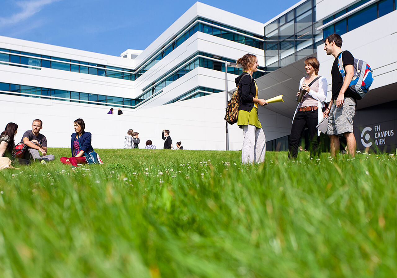 Gebäude der FH Wien mit Studierenden draußen auf einer Wiese vor den weißen Gebäuden.