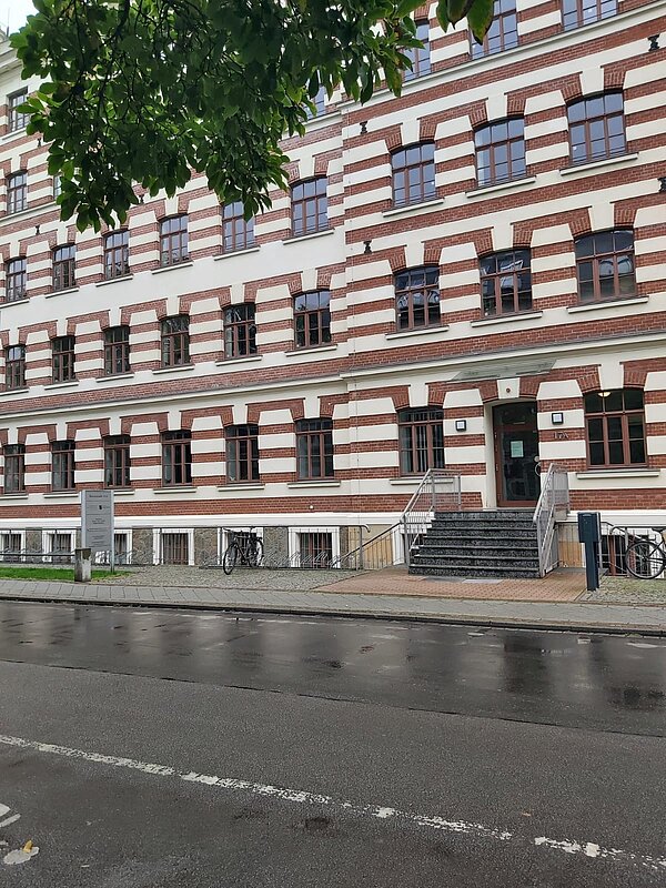 Das Bild zeigt die Regionalstelle Leipzig des Landesamtes für Schule und Bildung. Das Gebäude hat eine auffällige Fassade mit rot-weißen Ziegelmustern und großen Fenstern, die symmetrisch angeordnet sind. Der Eingangsbereich ist durch eine Treppe erreichbar, die zu einer Tür mit einem kleinen Vordach führt. Rechts und links vom Eingang stehen Fahrradständer.
