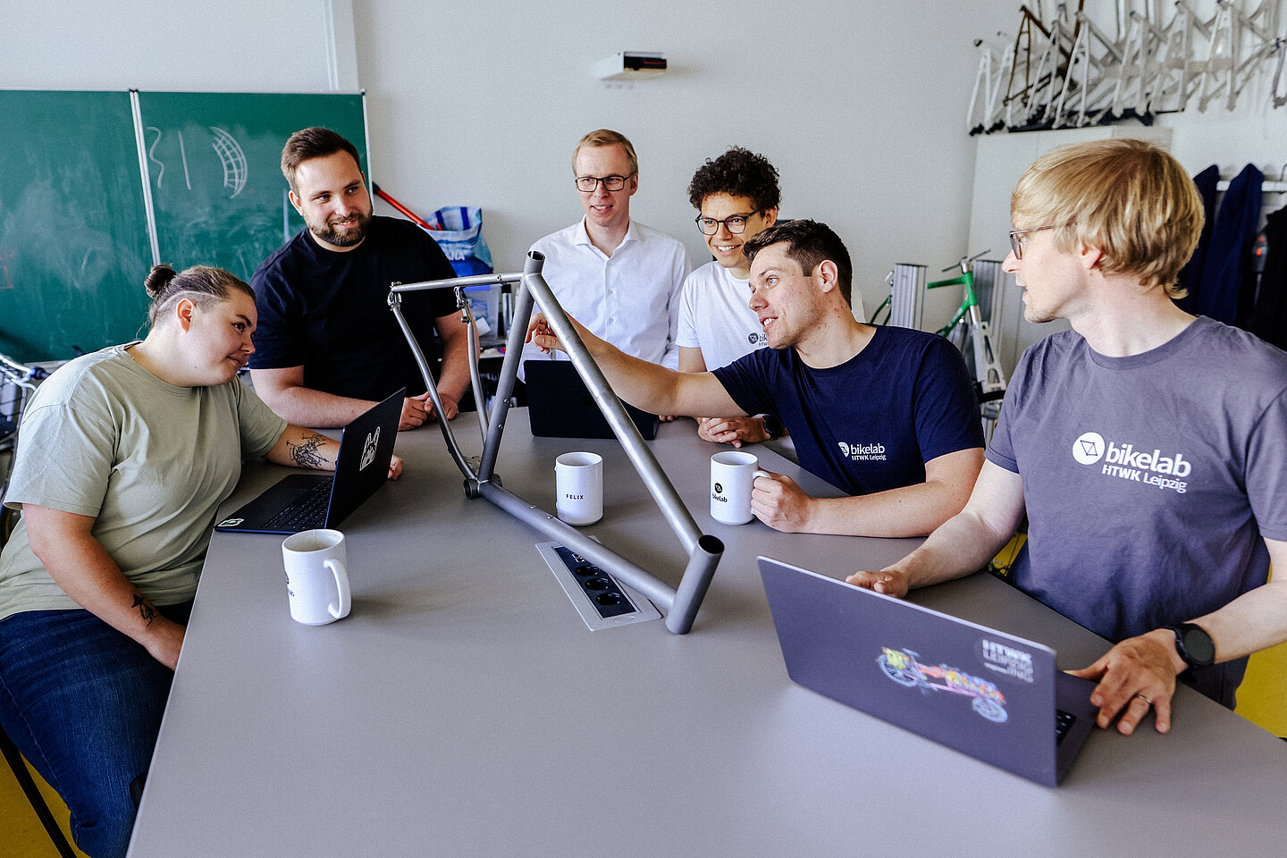 Sechs Personen sitzen um einen Tisch, betrachten gemeinsam einen Fahrradrahmen, Laptops und Tassen vor sich.