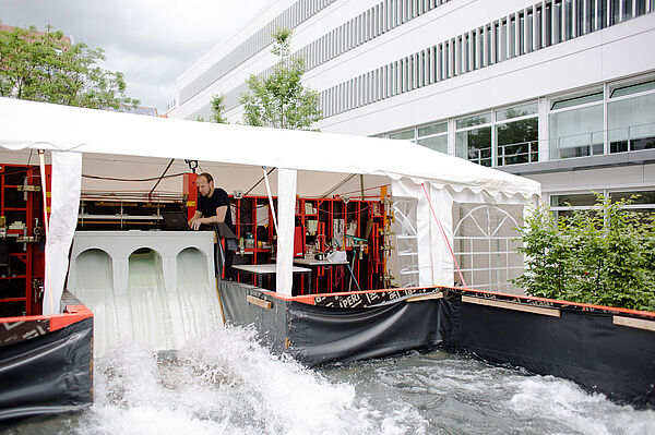 Unter einem langen Pavillon sind im Freien viele Geräte des Wasserbaulabors aufgebaut. In der Mitte ist eine Talsperre errichtet, durch die Wasser fließt