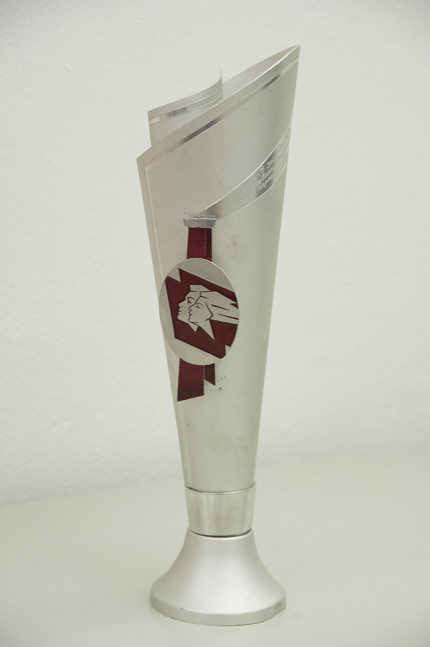Pokal aus dem HTWK-Archiv (Foto: HTWK Leipzig) Zu sehen ist ein silberner Pokal, der fackelförmig gebaut ist. An seiner Seite hängt an einem kurzen roten Band eine runde, flache Scheibe, auf der die Köpfe eines Mannes und einer Frau abgebildet sind. Beide schauen nach links aus dem Bild und recken das Kinn leicht nach oben, die Haare der Frau wehen scheinbar im Wind.