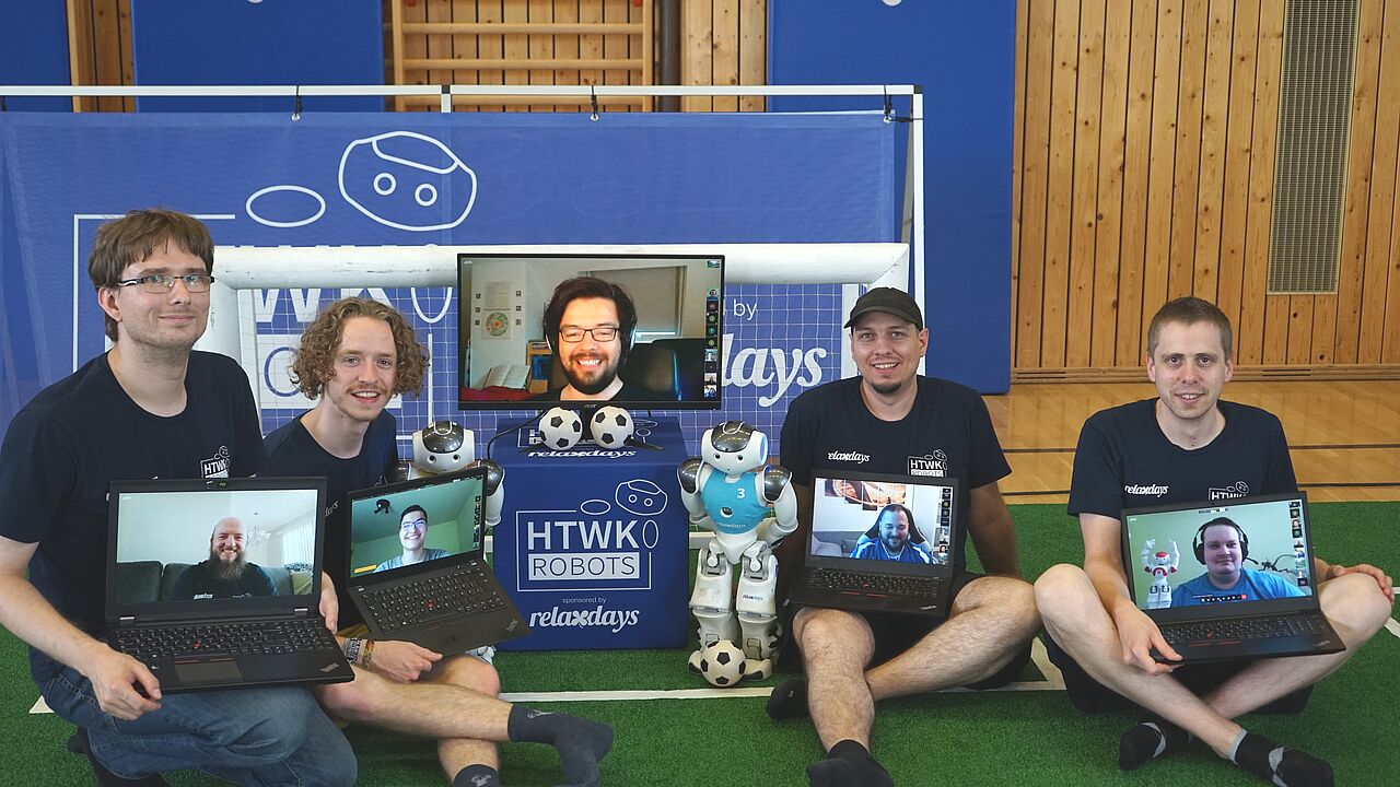 Vier Männer mit Laptops auf dem Schoß vor kleinem Tor und mit humanoidem Roboter