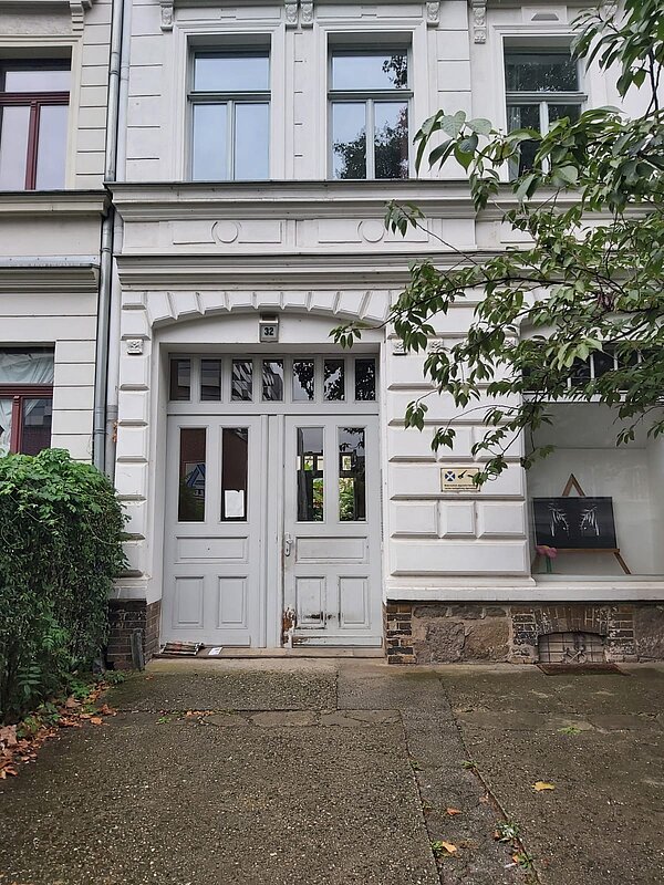 Eingang eines historischen Gebäudes mit einer weißen Fassade. Die Haustür ist zweiflügelig, aus Holz und in einem leicht abgenutzten Zustand. Oberhalb der Tür ist die Hausnummer „32“ angebracht.