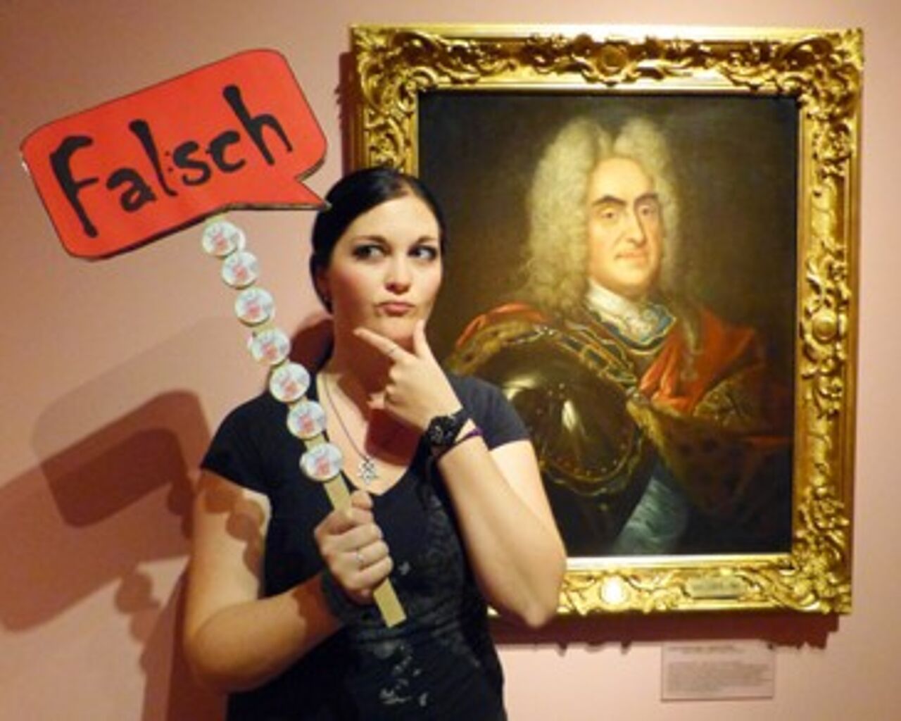Eine Absolventin des Studiengangs Museologie an der HTWK Leipzig steht in einer Ausstellung neben einem Gemälde von August dem Starken. Sie hält ein Schild mit der Aufschrit: „Falsch“, hoch. Sie greift sich nachdenklich ans Kinn.