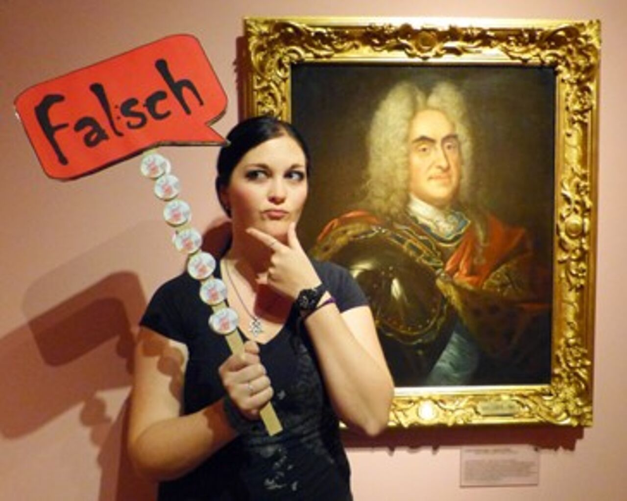 Gaumnitz_Franziska Eine Absolventin des Studiengangs Museologie an der HTWK Leipzig steht in einer Ausstellung neben einem Gemälde von August dem Starken. Sie hält ein Schild mit der Aufschrit: „Falsch“, hoch. Sie greift sich nachdenklich ans Kinn.