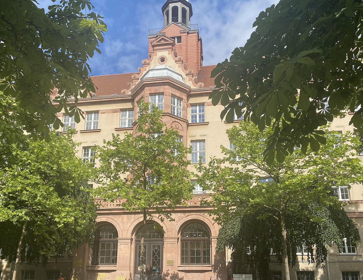 Das Bild zeigt ein großes, mehrstöckiges Gebäude mit beiger Fassade und roten Backsteinakzenten. Es hat ein rotes Ziegeldach, und im zentralen Bereich befindet sich ein kleiner Turm. Die Fenster sind symmetrisch angeordnet, und der Eingang ist von großen Bögen umrahmt. Um das Gebäude herum wachsen grüne Bäume, die den unteren Teil teilweise verdecken.