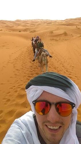 Sebastian Ehrhart reitet in der Wüste auf einem Kamel und schießt das Foto während des Ritts von sich selbst. Im Hintergrund sieht man eine Karawane aus weiteren Reitern.
