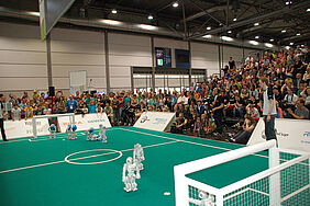 Die NAO-Roboter spielen auf einem Feld, die umliegenden Tribünen sind voll mit menschlichen Zuschauern