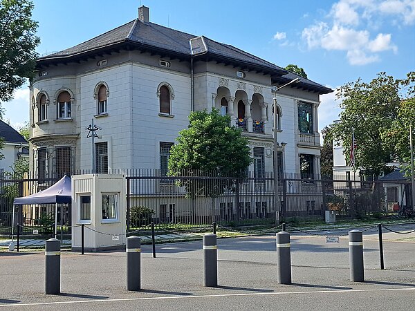 Ein beeindruckendes Altbaugebäude mit weißer Fassade und verzierten Rundbogenfenstern. Das Gebäude hat mehrere Balkone, dessen Geländer ebenfalls verziert sind. Das Gebäude ist von einem Zaun umgeben und befindet sich an einer Straße.
