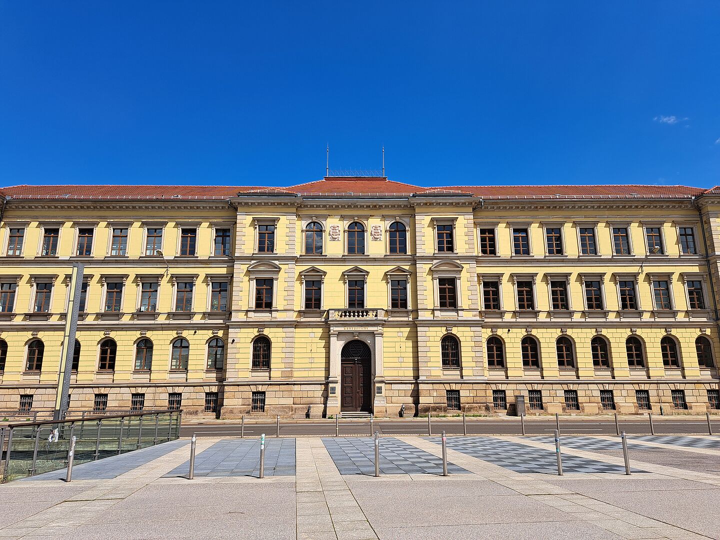 Das Bild zeigt das Landgericht Leipzig, ein historisches Gebäude mit einer beeindruckenden gelben Fassade. Es zeichnet sich durch symmetrische Fensterreihen und zentrale Eingangstüren aus. Die Architektur ist geprägt durch klassische Säulen und detaillierte Friesverzierungen oberhalb der Fenster. Das Gebäude hat ein rotes Ziegeldach und die Fassade ist klar und gut erhalten.
