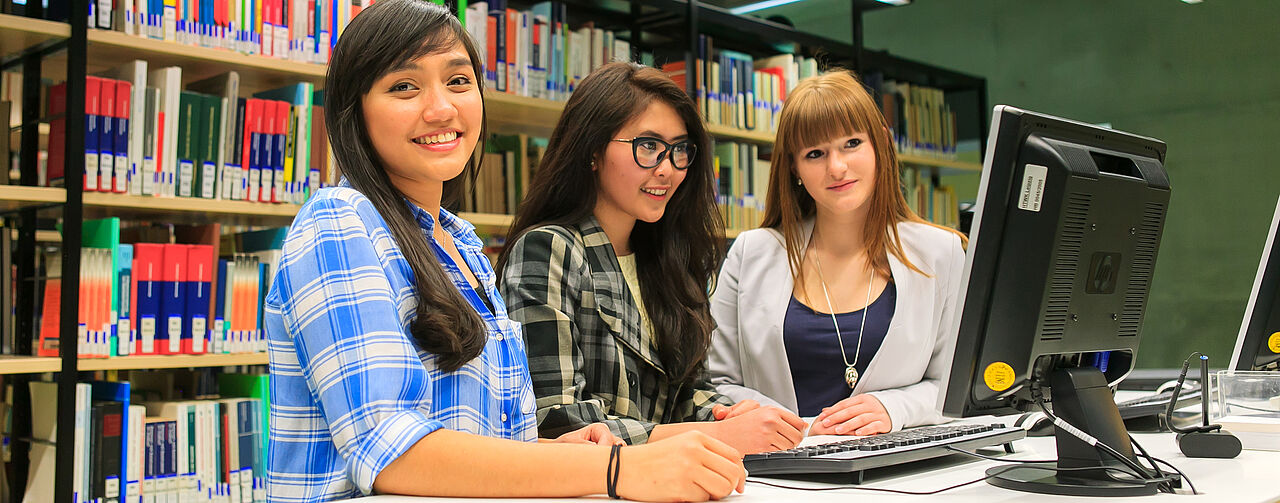 Three students are standing in the library in front of a computer