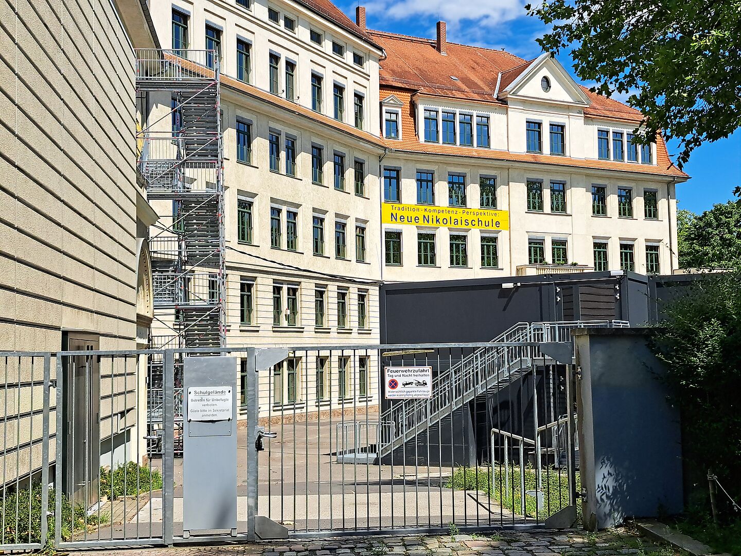 Das Bild zeigt ein großes, vierstöckiges Schulgebäude der "Neue Nikolaischule" mit traditioneller Architektur, einschließlich einem Giebeldach und symmetrisch angeordneten Fenstern. Das Gebäude ist in hellen Farbtönen gehalten. Im Vordergrund ist ein graues Metalltor zu sehen, das Zugang zum Schulgelände gewährt. Rechts neben dem Tor befindet sich eine Außentreppe, die zu einem Conatinergebäude führt. Der Himmel ist blau und der Vordergrund zeigt einige grüne Bäume, die teilweise das Gebäude verdecken.