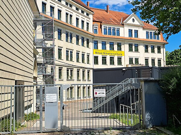 Das Bild zeigt ein großes, vierstöckiges Schulgebäude der "Neue Nikolaischule" mit traditioneller Architektur, einschließlich einem Giebeldach und symmetrisch angeordneten Fenstern. Das Gebäude ist in hellen Farbtönen gehalten. Im Vordergrund ist ein graues Metalltor zu sehen, das Zugang zum Schulgelände gewährt. Rechts neben dem Tor befindet sich eine Außentreppe, die zu einem Conatinergebäude führt. Der Himmel ist blau und der Vordergrund zeigt einige grüne Bäume, die teilweise das Gebäude verdecken.