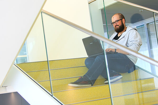 Tom Sperschneider sitzt auf einer Treppe im Uni-Gebäude mit einem Laptop auf den Knien.