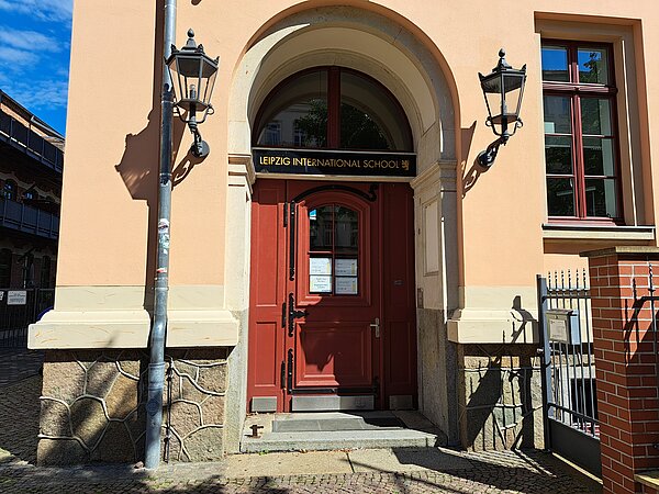 Das Bild zeigt den Eingang zur Internationalen Schule Leipzig. Der Eingangsbereich wird von einer hohen Wölbung geschmückt, die innen mit weißen Stuck ausgefüllt ist. Somit ist die Tür leicht in das Gebäude eingelassen, welhalb zwei steinerne Stufen zu ihr hinauf führen. Die Tür besteht aus braun-rotem Holz, ebenso wie die beiden Holbretter um sie herum. Der Türknauf ist golden und in der oberen Türhälfte befindet sich eine viergeteilte Scheibe, welche viereckig mit einer Wölbung der oberen Seite ist. Über der Tür ist ein goldener schriftzug auf schwarzem Stein gesetzt. Er trägt die Aufschrift "Leipzig International School" in Großbuchstaben. Darüber befindet sich eine weitere Glasscheibe in Form eines Halbkreises, welche von einem hölzernen Balken in zwei gesteilt wird. Rechts neben dem Eingang ist ein rechteckiges Fenster sowie eine Backsteinmauer mit einem Eisentor zu sehen. Das Gebäude selbst ist gremfarben gestrichen und unten an der Fassade mit Steinplatten beschmückt. Links und rechts neben der Eingangstür befinden sich zwei Laternen, im selben Stil wie das Tor gehalten. Sie sind auf der Höhe des Schriftzuges angebracht.
