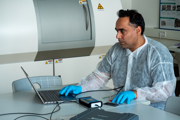 Wissenschaftler sitzt am Schreibtisch vor Laptop und dem Analysegerät in der Hand, hellblaue Gummi-Handschuhe sowie Ganzkörperschutzanzug aus dünnem Fleck