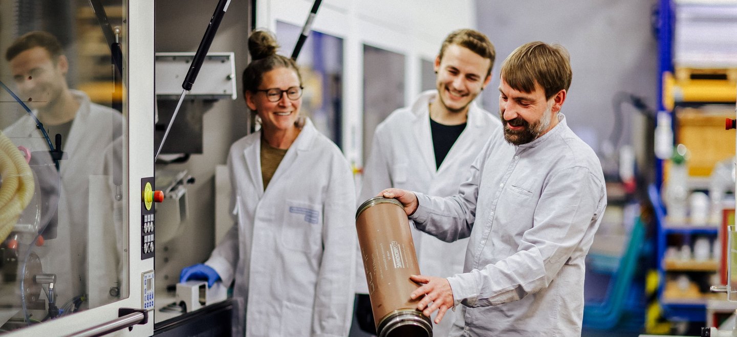 Drei Personen, zwei im Laborkittel, begutachten gemeinsam einen Druckzylinder vor einer Beschichtungsanlage.